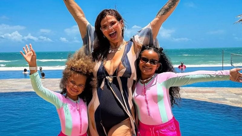 Thais Carla com as duas filhas crianças posando sorridentes na praia com roupas de banho coloridas, braços abertos e fundo de mar e céu azul em dia de sol