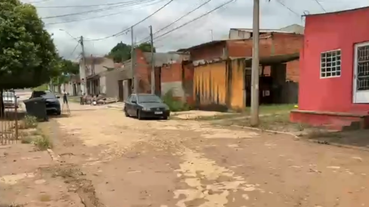 Rua de Capivari com casas de cores variadas, terreno de chão batido, carros estacionados e poste de iluminação ao fundo, em uma área urbana.
