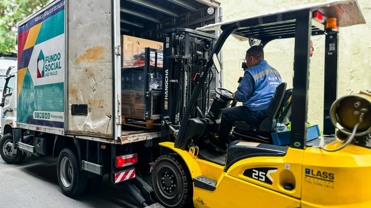 Empilhadeira amarela operada por trabalhador ao lado de caminhão de carga, com caixas e equipamentos ao fundo, em ambiente externo.