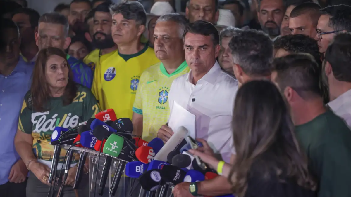 Imagem de coletiva de imprensa com um homem de camisa branca, cercado por jornalistas e microfones em um ambiente lotado de público e apoiadores.
