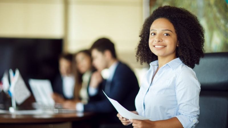 Servidora pública em ambiente de escritório, sentada em sala de atendimento, sorrindo e segurando documentos enquanto colegas trabalham ao fundo.