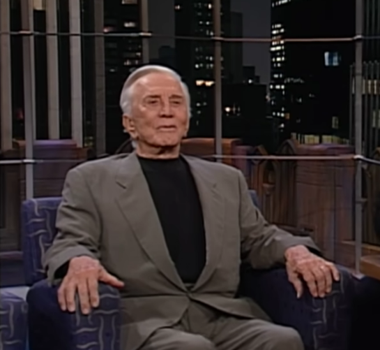Kirk Douglas em entrevista em um cenário urbano à noite, sentado em uma poltrona, com prédios iluminados ao fundo.
