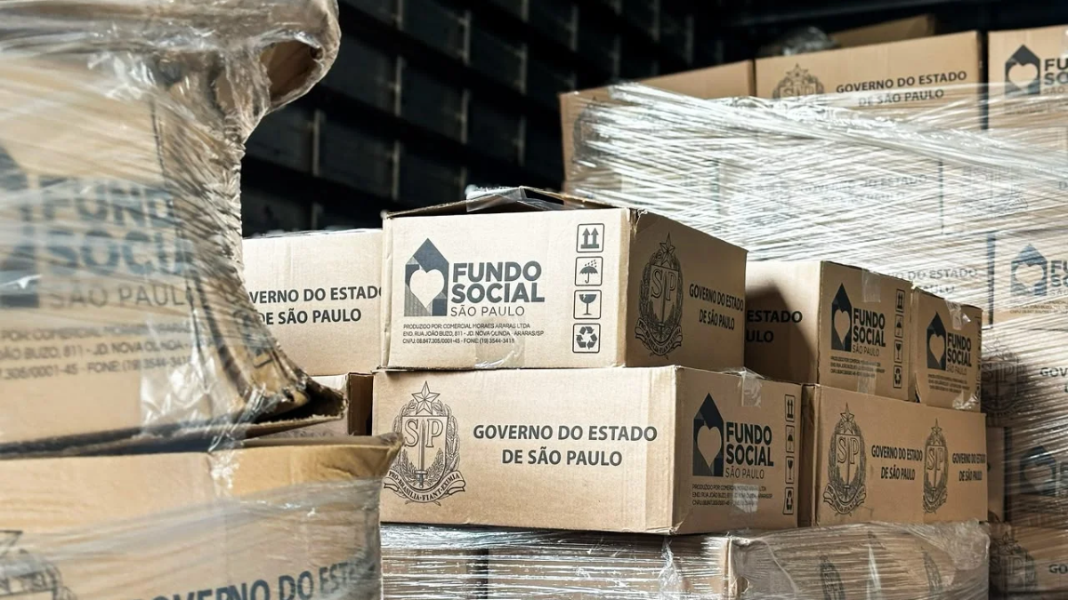 Caixas de papelão com o logotipo “Fundo Social São Paulo” e “Governo do Estado de São Paulo” armazenadas em um depósito, com fardos embalados ao fundo.