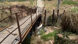 Ponte de concreto com trilhos enferrujados e cercas metálicas em área rural, com valeta e vegetação ao redor, vista do interior de um antigo desvio