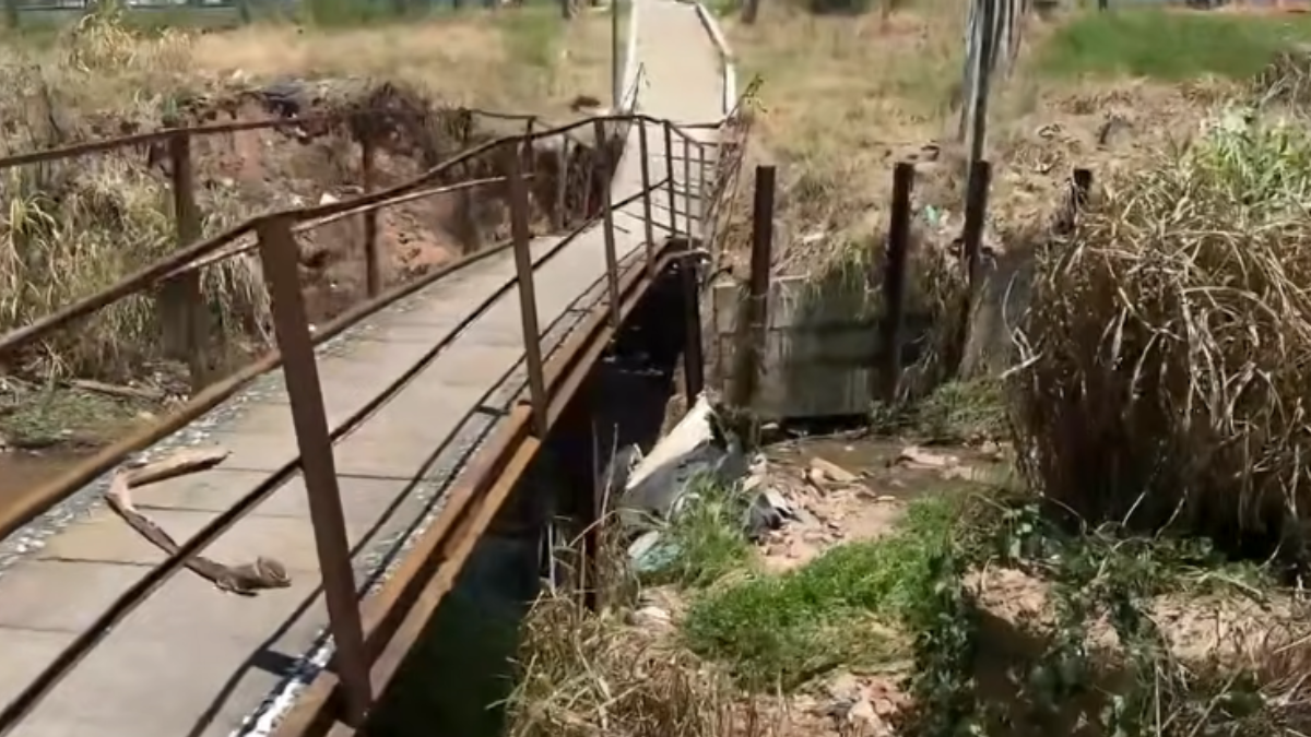 Ponte de concreto com trilhos enferrujados e cercas metálicas em área rural, com valeta e vegetação ao redor, vista do interior de um antigo desvio