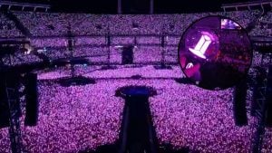 Montagem de show em estádio lotado com pessoas segurando objetos brilhantes e um lightstick iluminado em tons de roxo e rosa
