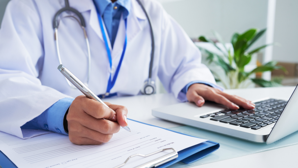 Médico em consultório preenchendo documentos com caneta e usando notebook em uma mesa, com estetoscópio ao redor do pescoço