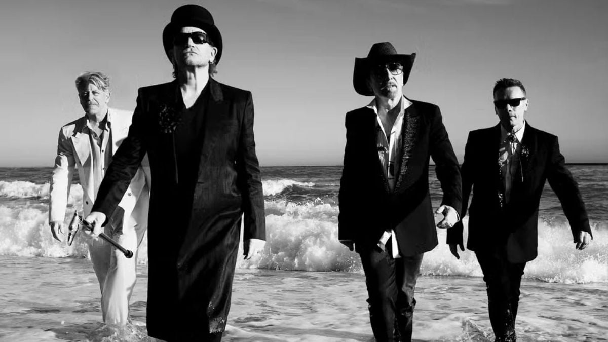Fotografia em preto e branco de quatro homens da banda U2 com chapéus e ternos, andando pela praia com ondas ao fundo, em estilo clássico de cinema.