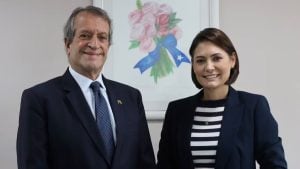Foto de Valdemar Costa Neto e Michelle Bolsonaro de terno em ambiente corporativo, sorrindo para a câmera, com quadro floral ao fundo. Mulher e homem posam com postura formal.