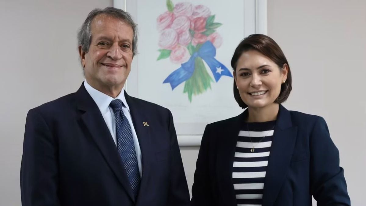 Foto de Valdemar Costa Neto e Michelle Bolsonaro de terno em ambiente corporativo, sorrindo para a câmera, com quadro floral ao fundo. Mulher e homem posam com postura formal.