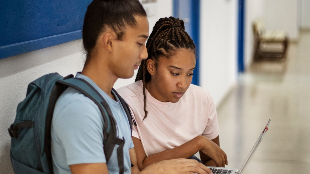 Três pessoas trabalhando em equipe em uma mesa, com a mulher de cabelo cacheado analisando um documento no tablet, enquanto os demais observam e conversam em um ambiente de escritório ensolarado.