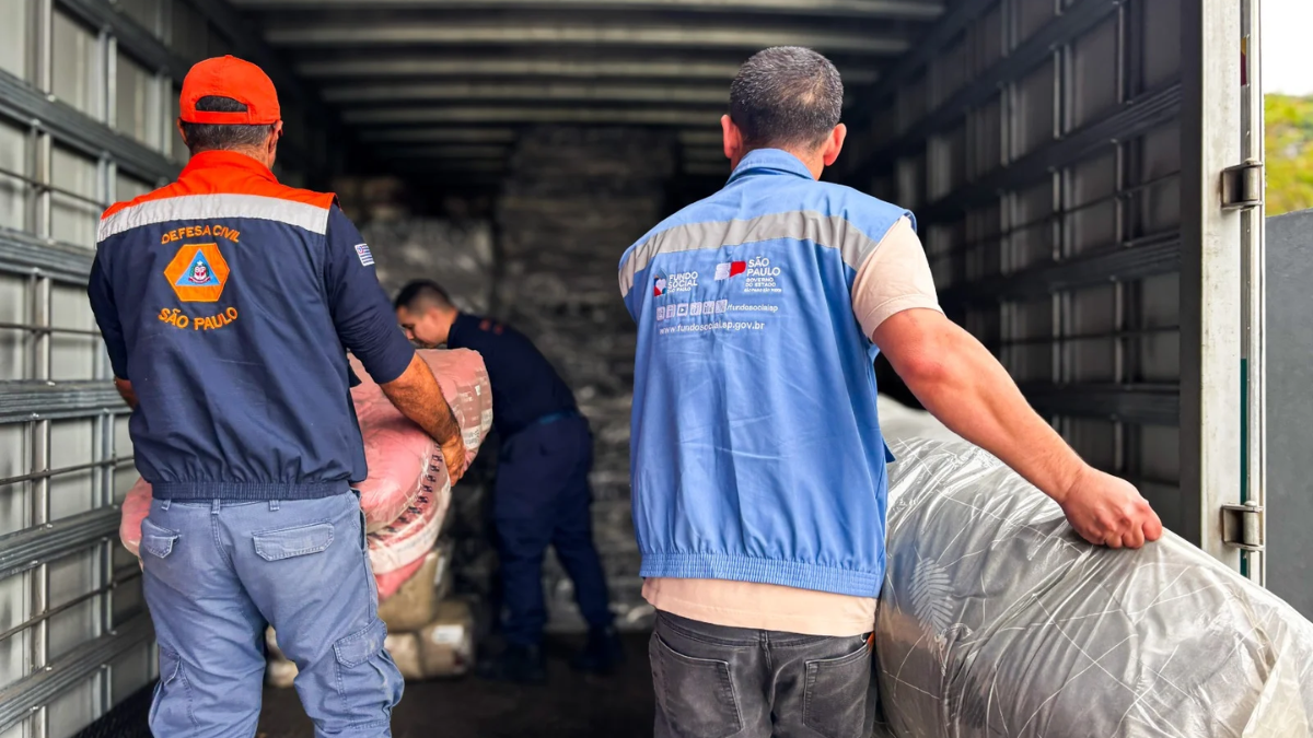 Trabalhadores com coletes e uniformes de empresa transportando cargas em um caminhão, com um entregador empurrando um volume coberto durante o carregamento.
