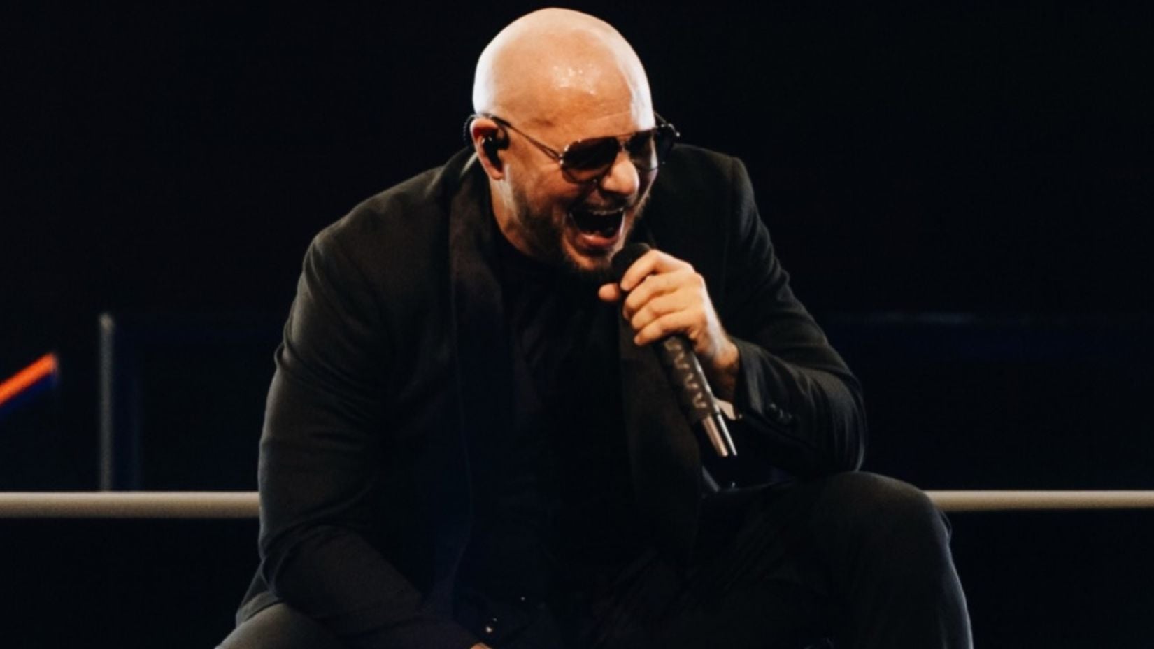 Pitbull em apresentação ao vivo, sentado em palco com microfone na mão, usando óculos escuros e jaqueta preta, em ambiente com luzes de show e fundo escuro.