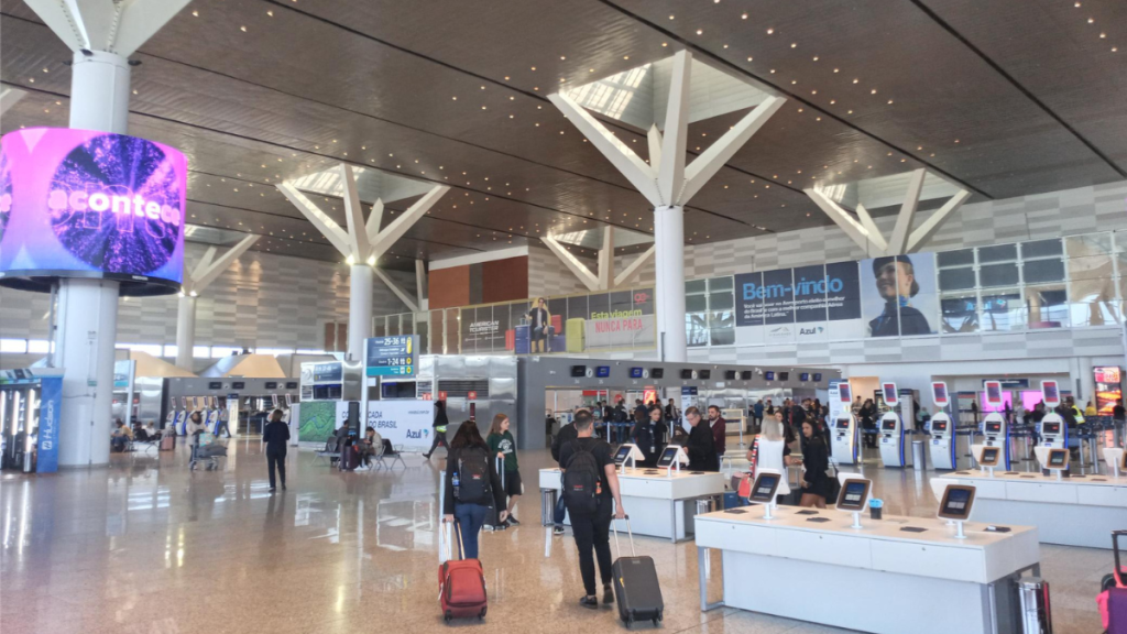 Interior de um aeroporto movimentado com balcões de atendimento, equipamentos de autoatendimento, placas de Bem-vindo e pessoas circulando no saguão sob iluminação de teto