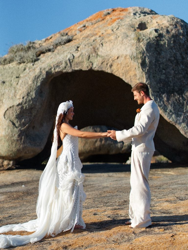 Casamento intimista de Juliette com Kaique Cerveny em Lajedo de Pai Mateus, na Paraíba: noivo e noiva em trajes brancos trocam um gesto de carinho ao ar livre diante das pedras