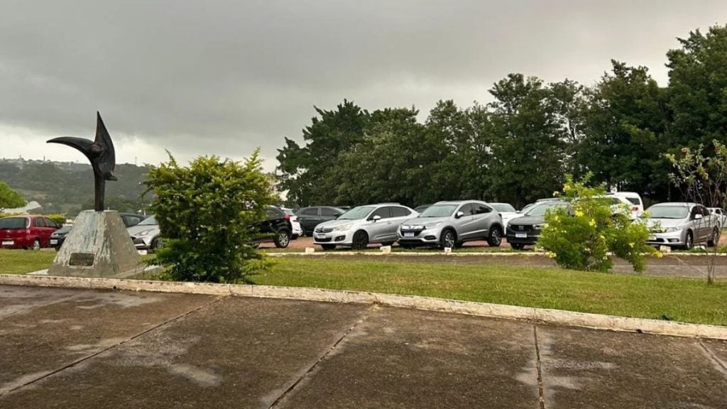 Área externa com estacionamento, jardins e monumento em cemitério de Campinas durante dia nublado.