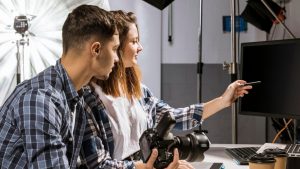 Estudio de fotografia, com fundo branco e cinza, equipamentos de luz. Homem e mulher apontam para o computador, com uma camera fotográfica na mão.
