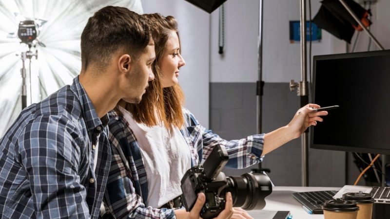 Estudio de fotografia, com fundo branco e cinza, equipamentos de luz. Homem e mulher apontam para o computador, com uma camera fotográfica na mão.