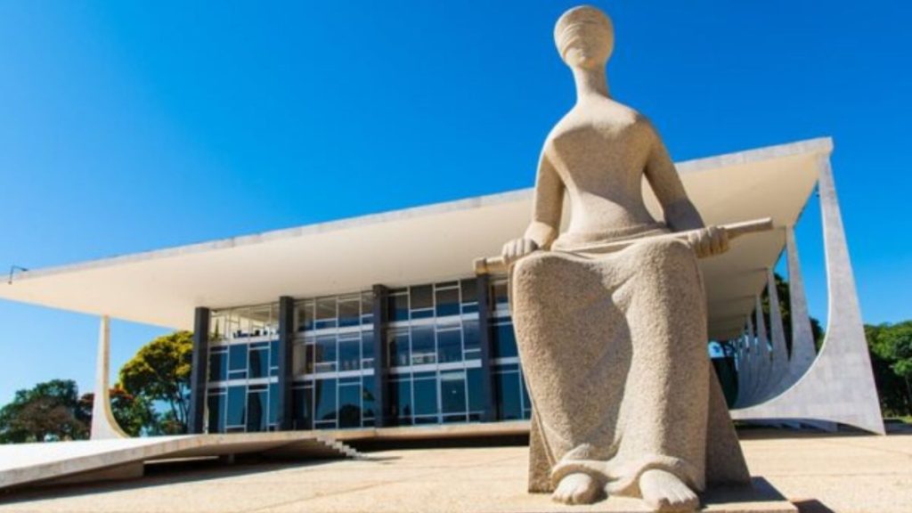 Vista frontal do edifício do Supremo Tribunal Federal (STF) em Brasília, com a escultura da Justiça em primeiro plano sob um céu azul límpido.