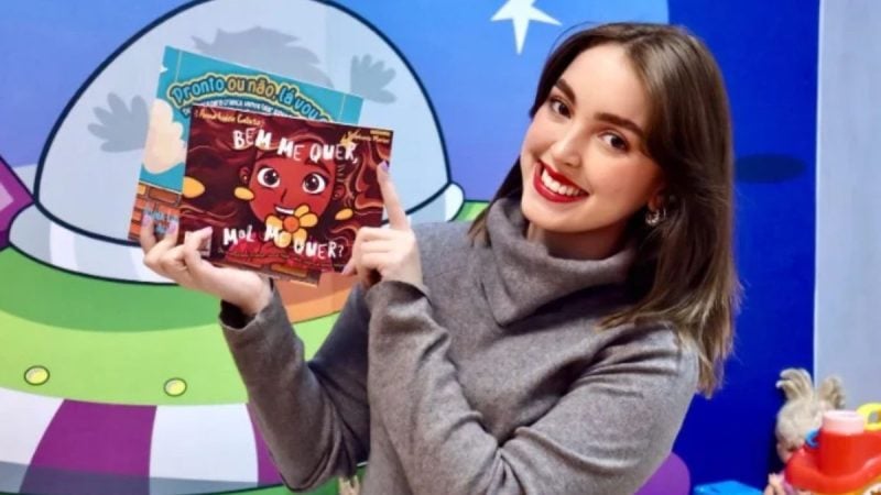Mulher sorridente em ambiente infantil segura um livro escrito “Bem Me Quer, Mal Me Quer” com ilustração de um personagem e aponta para a embalagem, em foto promocional.