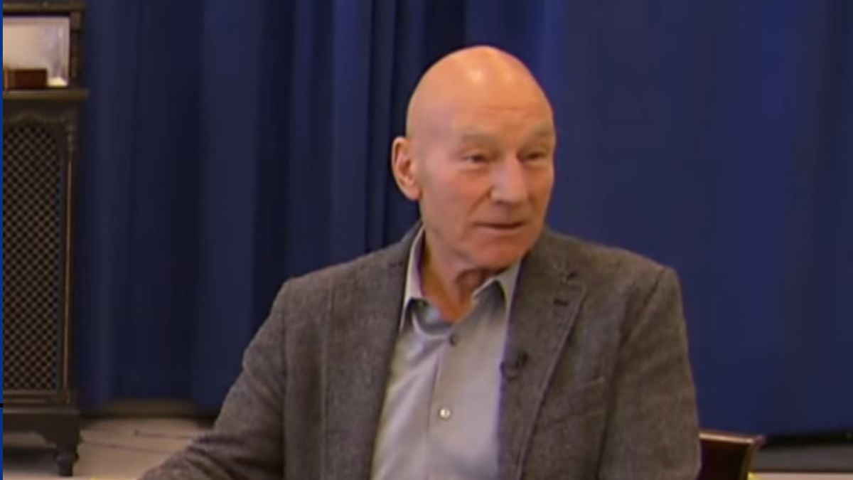 Patrick Stewart em uma entrevista, sentado em um palco com fundo azul e blusa social clara, expressando atenção durante a fala.
