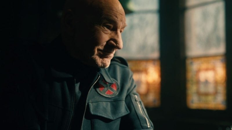 Patrick Stewart em um ambiente interno, usando jaqueta escura e encarando para o lado com expressão séria, em plano próximo e iluminação suave.